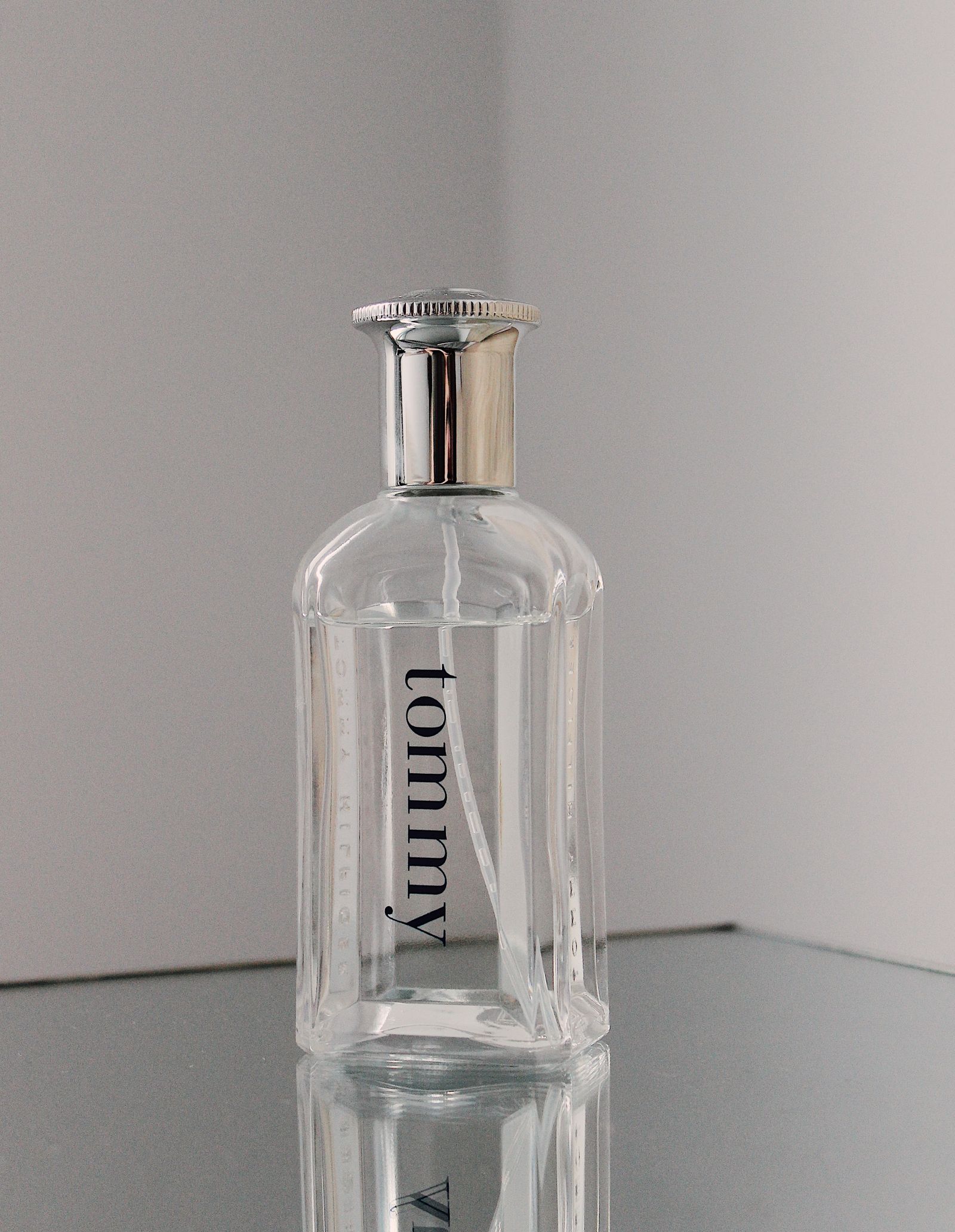 Original perfume photograph: clear glass bottle, silver cap, vertical tommy lettering and a reflection on a mirrored surface.