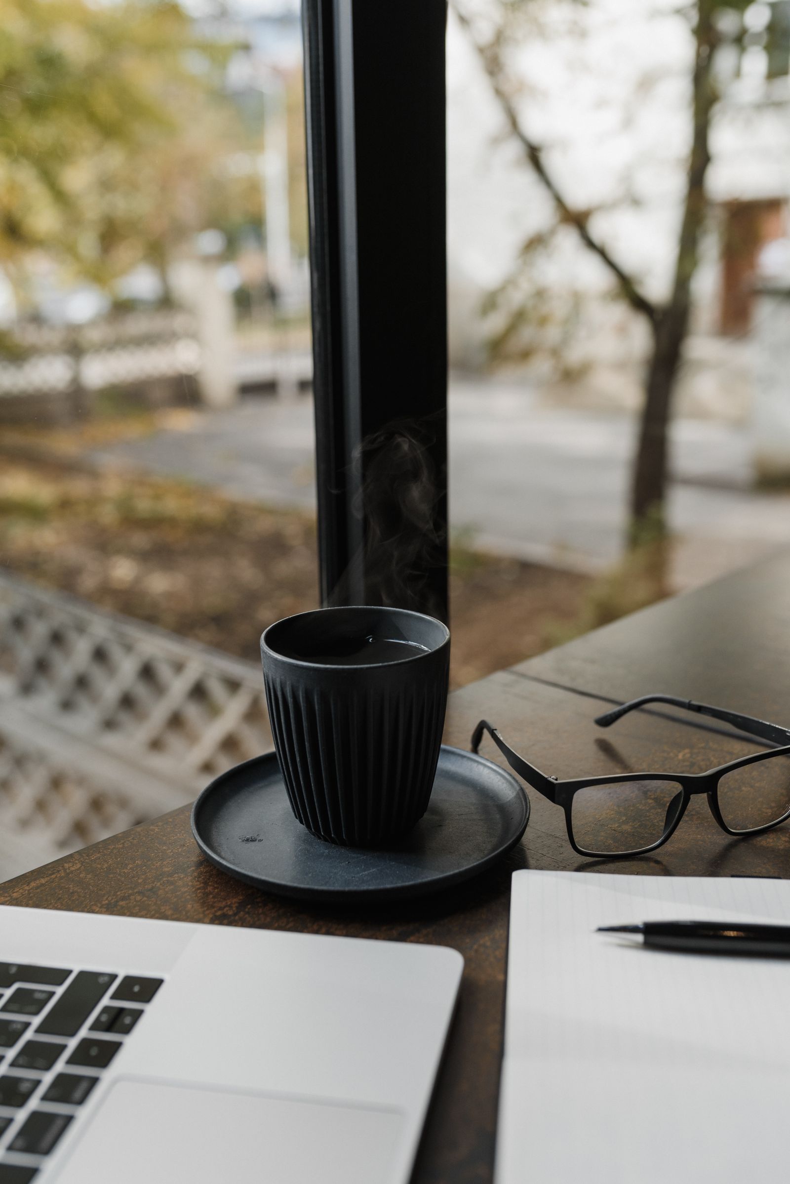 Original cluttered desk photo with a black cup and saucer, glasses, laptop and notebook